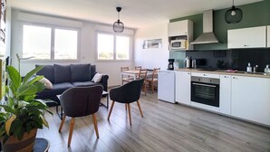 Cocina moderna con gabinetes blancos, microondas y cafetera. Cuenta con un comedor con mesa y sillas, y una sala de estar con sofá y planta.