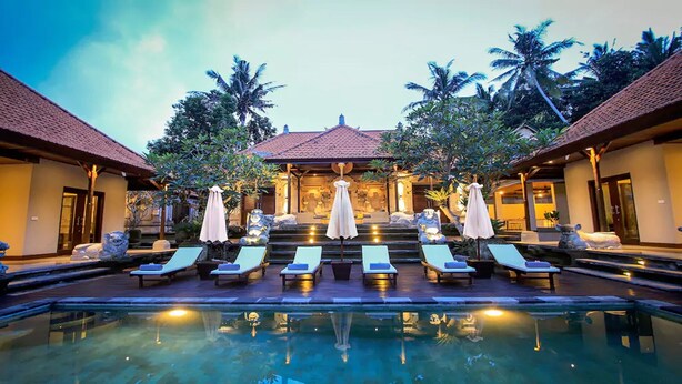 A poolside area with lounge chairs and umbrellas, a stone statue, and a building with a tiled roof in the background. - Bali style villa 5 minutes from ubud center! (Ubud)