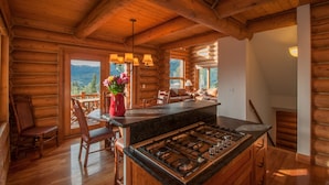 A log cabin interior with a kitchen area featuring a central island with a stove, wooden dining chairs, and a view of the mountains through large windows.