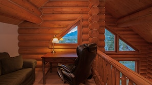 A log cabin interior with a wooden ceiling, a desk with a lamp, a leather chair, a green sofa, and a view of mountains through a window.