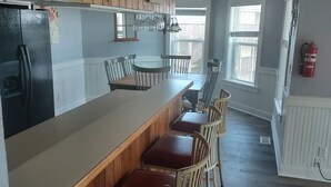 Cocina con armarios de madera, un refrigerador y una barra con taburetes. Se ven ventanas y un extintor.
