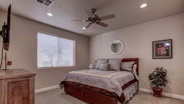 A bedroom with a bed, a ceiling fan, a window with blinds, a bedside table, and a potted plant.
