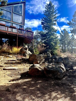 A modern building with large windows and a wooden deck, surrounded by rocky terrain and pine trees.
