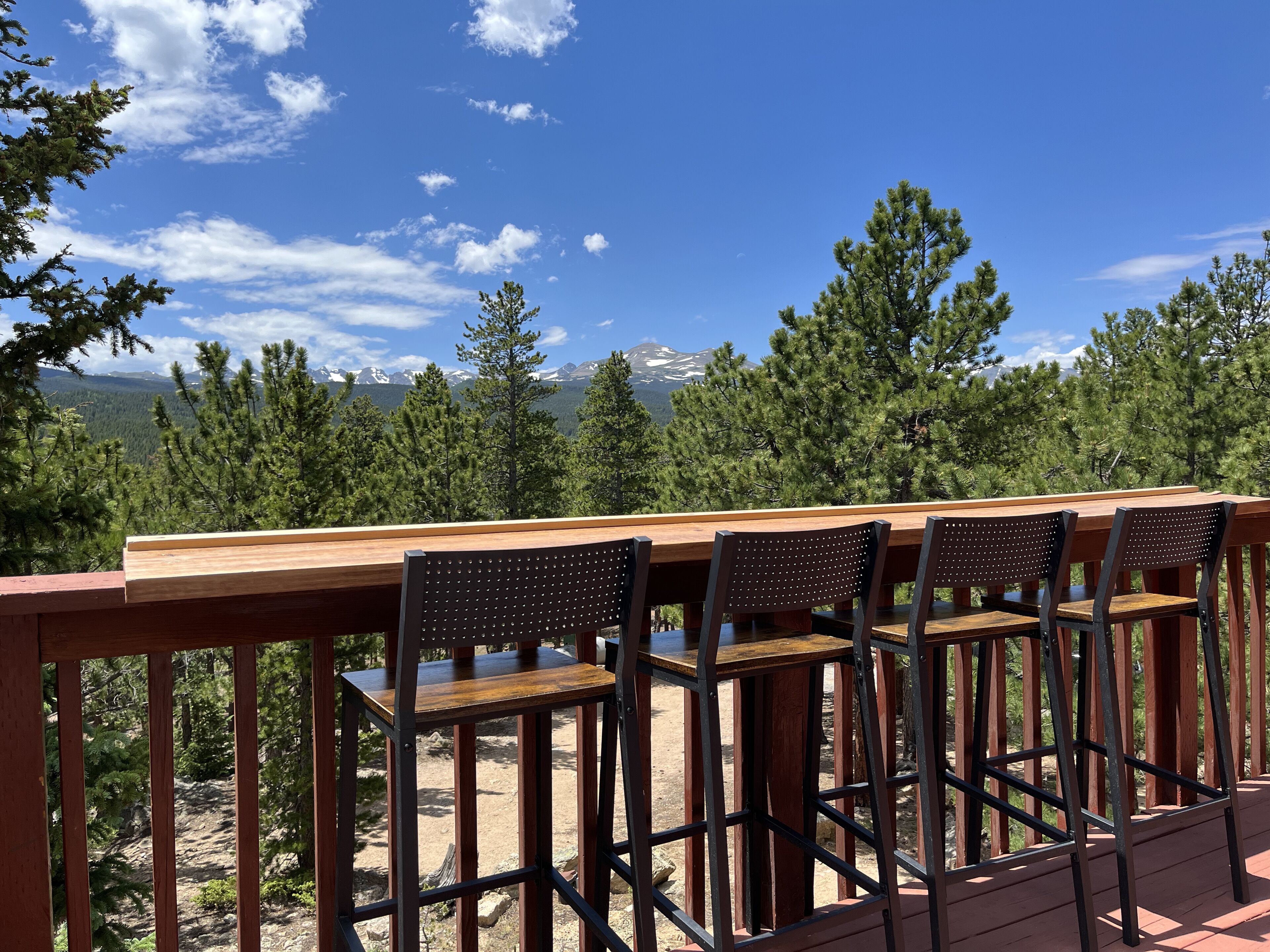 Una terraza de madera con una mesa y cuatro sillas, con vistas a un paisaje montañoso con árboles y cielo despejado.