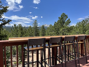 A wooden deck with a table and four chairs, overlooking a mountainous landscape with trees and a clear sky.