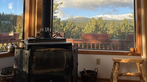 A rustic cabin interior with a large wood-burning stove, wooden furniture, and a view of a forested mountain range through large windows. - Stunning Views Of The Indian Peaks On Mountain Lake (Ward)