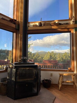 A rustic cabin interior with a large wood-burning stove, wooden furniture, and a view of a forested mountain range through large windows.
