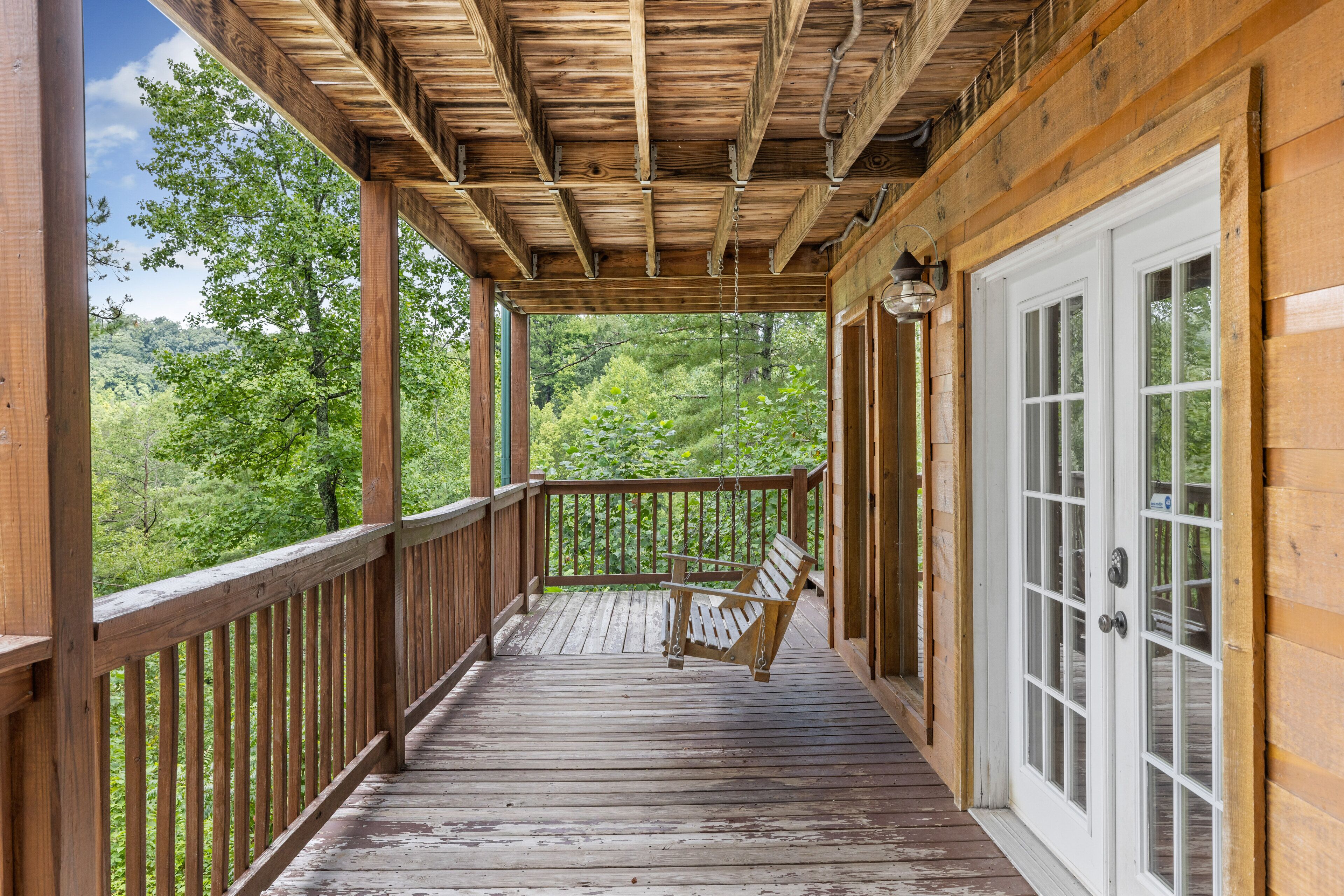 A wooden porch with a swing chair, a white door, and a view of a forested area.