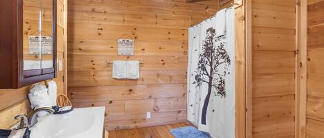 Un bagno in una baita di legno, con un lavandino bianco, uno specchio, un asciugamano appeso al muro e una tenda della doccia con un motivo alberato.
