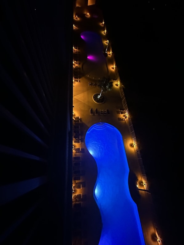 Vista aérea de um hotel com uma estrutura azulada e imponente que se assemelha a uma figura humana, cercada por quartos uniformemente espaçados e uma área circular central com uma fonte.