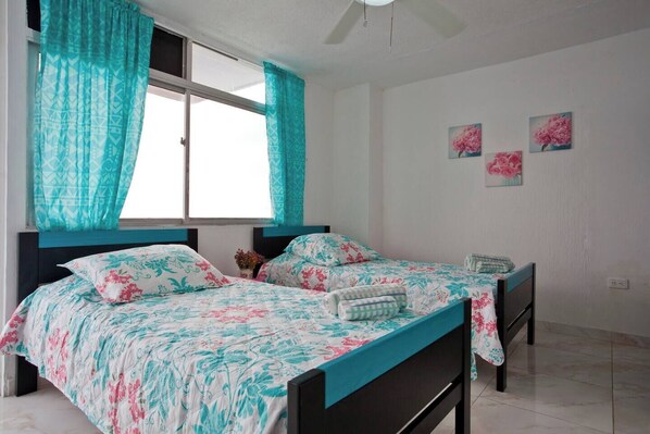 Une chambre avec deux lits, des rideaux turquoise, une literie à motifs floraux et un sol carrelé de couleur blanche.