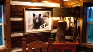 Ein rustikales Blockhaus-Interieur mit einem Holzstuhl, einem Bärenbild in einem Rahmen, einer Lampe und Ski, die an der Wand lehnen.