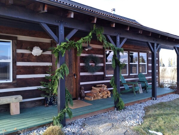 Une cabane en rondins dotée d’un porche décoré de plantes vertes et d’une guirlande. On y trouve une pile de bois de chauffage et un banc.