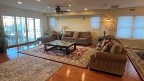 A spacious living room with a ceiling fan, large windows, and a sliding glass door leading to a balcony. - SPACIOUS HOUSE WITH POOL OCEANVIEW 3 DECKS 4 MASTER CLOSE TO OCEAN AND BOARDWALK (Wildwood Crest)