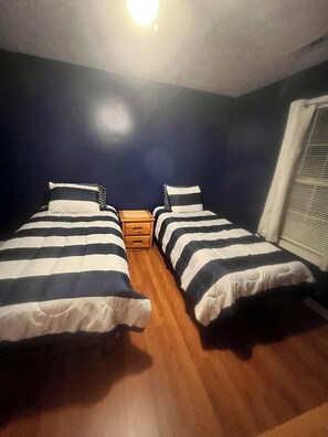 A room with two beds, a wooden nightstand, and a window with blinds.