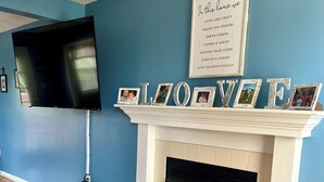 Un soggiorno con una parete blu, una TV a schermo piatto fissata al muro, un caminetto bianco decorato con cornici e foto, e un cartello incorniciato sopra il caminetto.