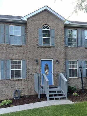 A two-story brick building with a blue door, grey shutters, and a small front porch with steps.
