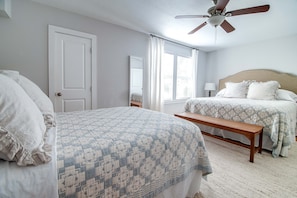 A bedroom with two beds, a ceiling fan, a bench, and a window with curtains.