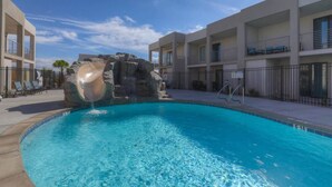 A swimming pool with a rock feature, adjacent to a building with balconies and a clear sky. - The Lofts #14 | Townhome Pool Resort | Poolside (Saint George)