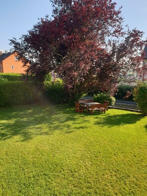 A well-maintained lawn with a wooden bench and a table, surrounded by hedges and a large tree with red leaves. - Family friendly accommodation (Furth im Wald)