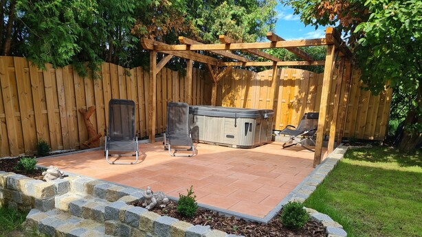 Une terrasse à l’arrière de la maison, avec un jacuzzi, deux chaises de jardin et une pergola en bois.