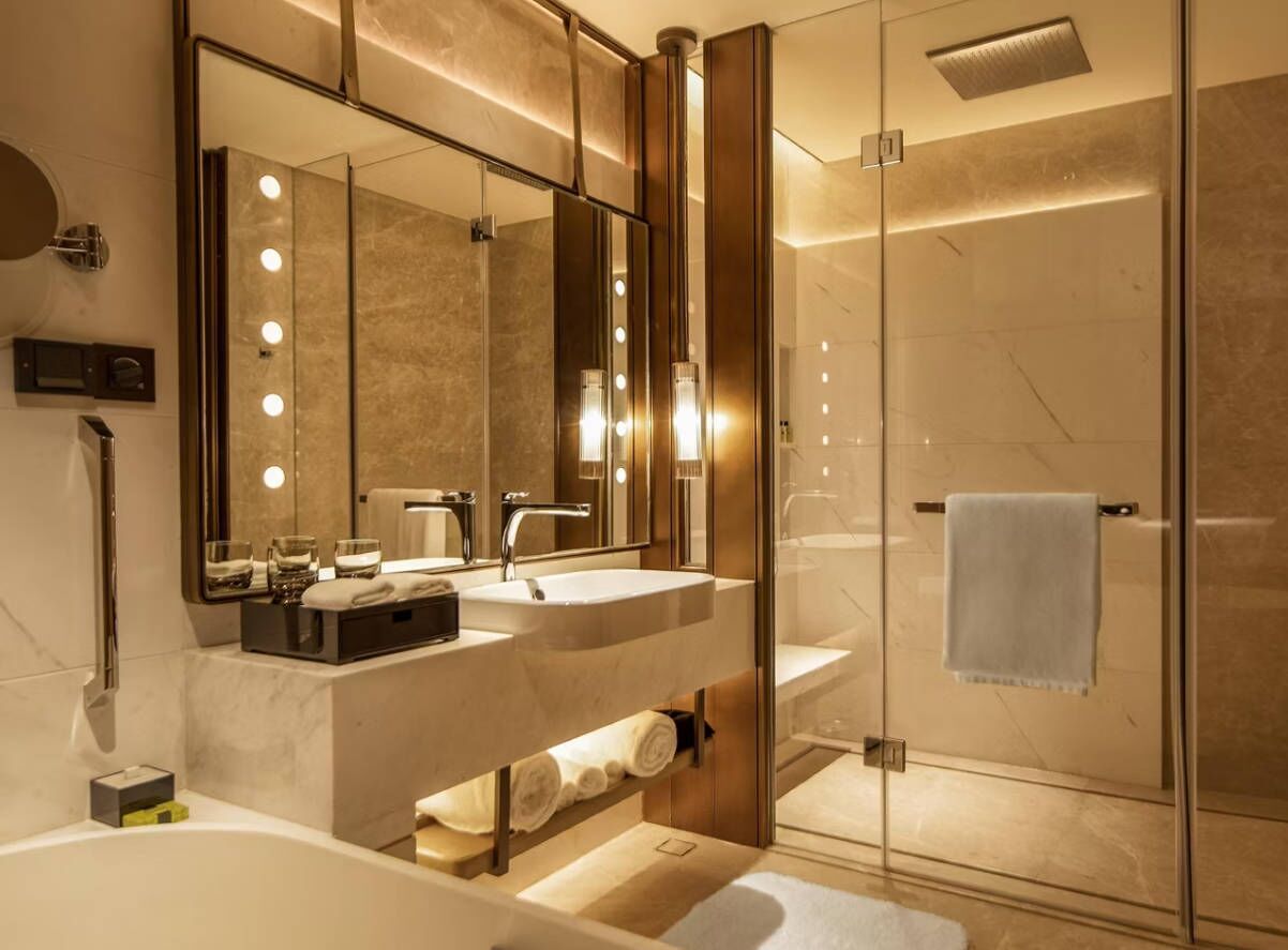 Ein modernes Badezimmer mit einer gläsernen Duschkabine, einem Waschtisch mit zwei Waschbecken und einem großen, beleuchteten Spiegel.