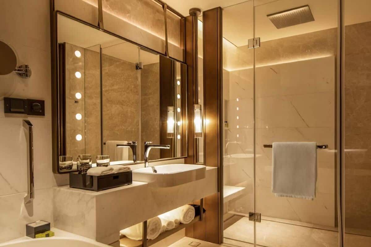 Un baño moderno con ducha envuelta en vidrio, tocador con dos lavamanos y un espejo grande con iluminación.
