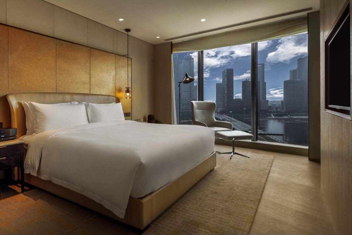 Habitación de hotel moderna con una cama grande, un escritorio con lámpara, una silla y vistas de la silueta urbana a través de amplias ventanas.