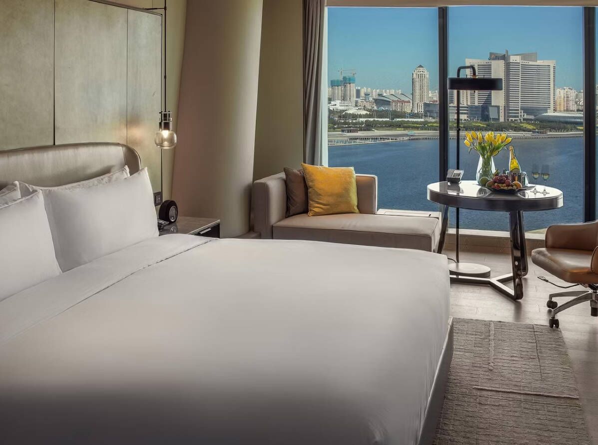 Ein Hotelzimmer mit einem großen Bett, einem Schlafsofa, einem kleinen Tisch mit einer Vase und Flaschen sowie Blick auf eine Stadtlandschaft und Wasser.