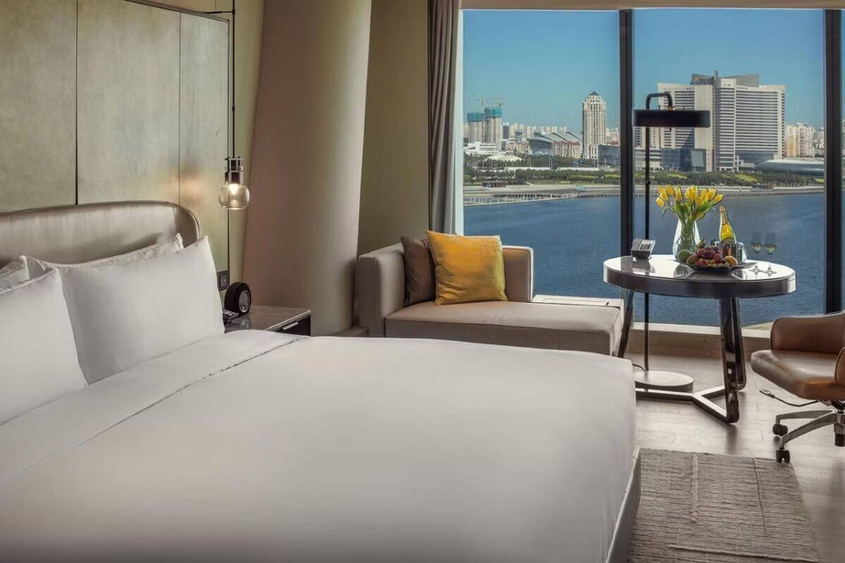 Habitación de hotel con una cama grande, un sofá, una mesita con un florero y botellas, y vistas a un horizonte urbano y al agua.