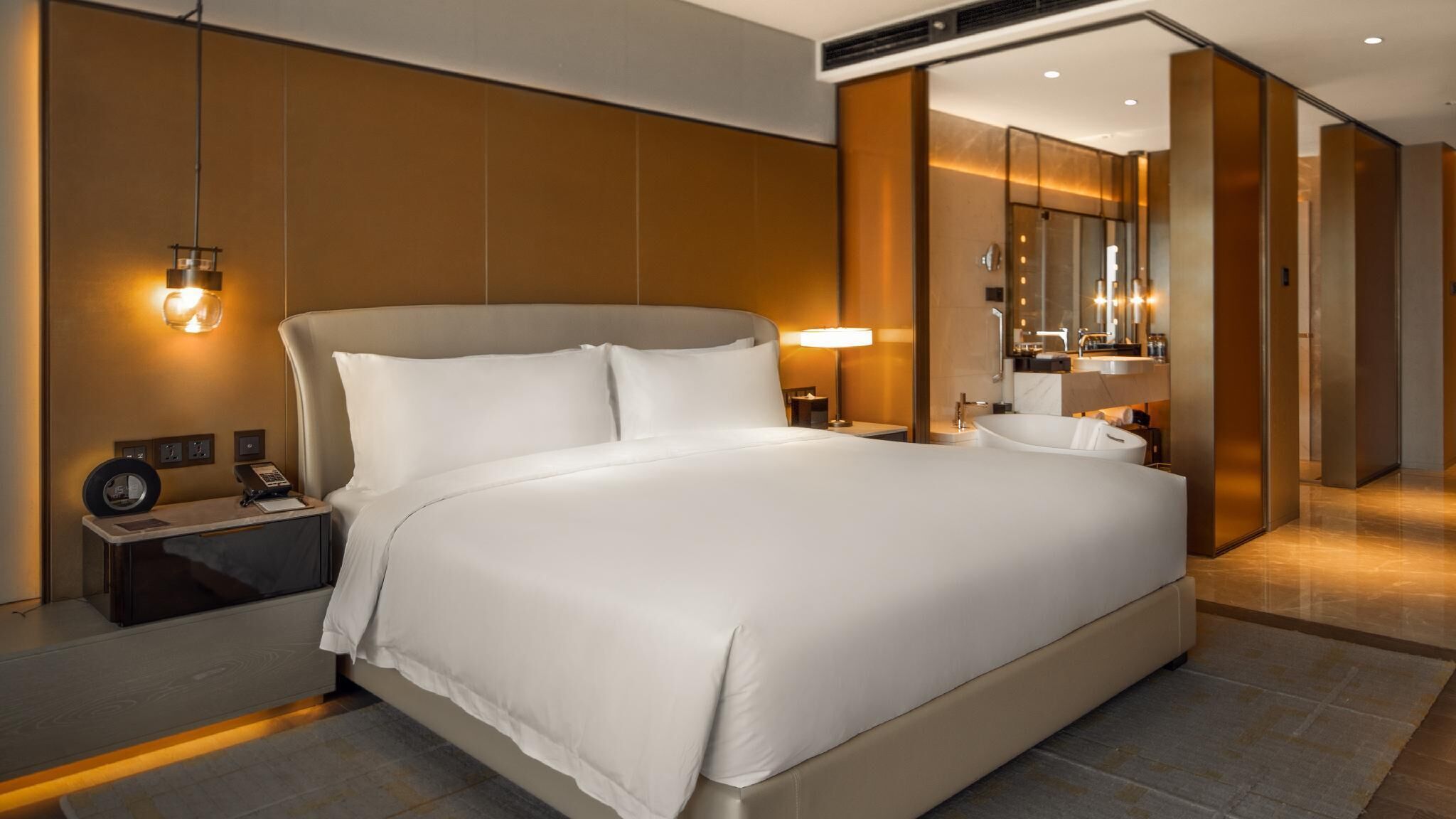 Ein modernes Hotelzimmer mit einem großen Bett, Nachttischlampen, einem Schreibtisch mit Telefon sowie einem Badezimmer mit Badewanne und Spiegel.