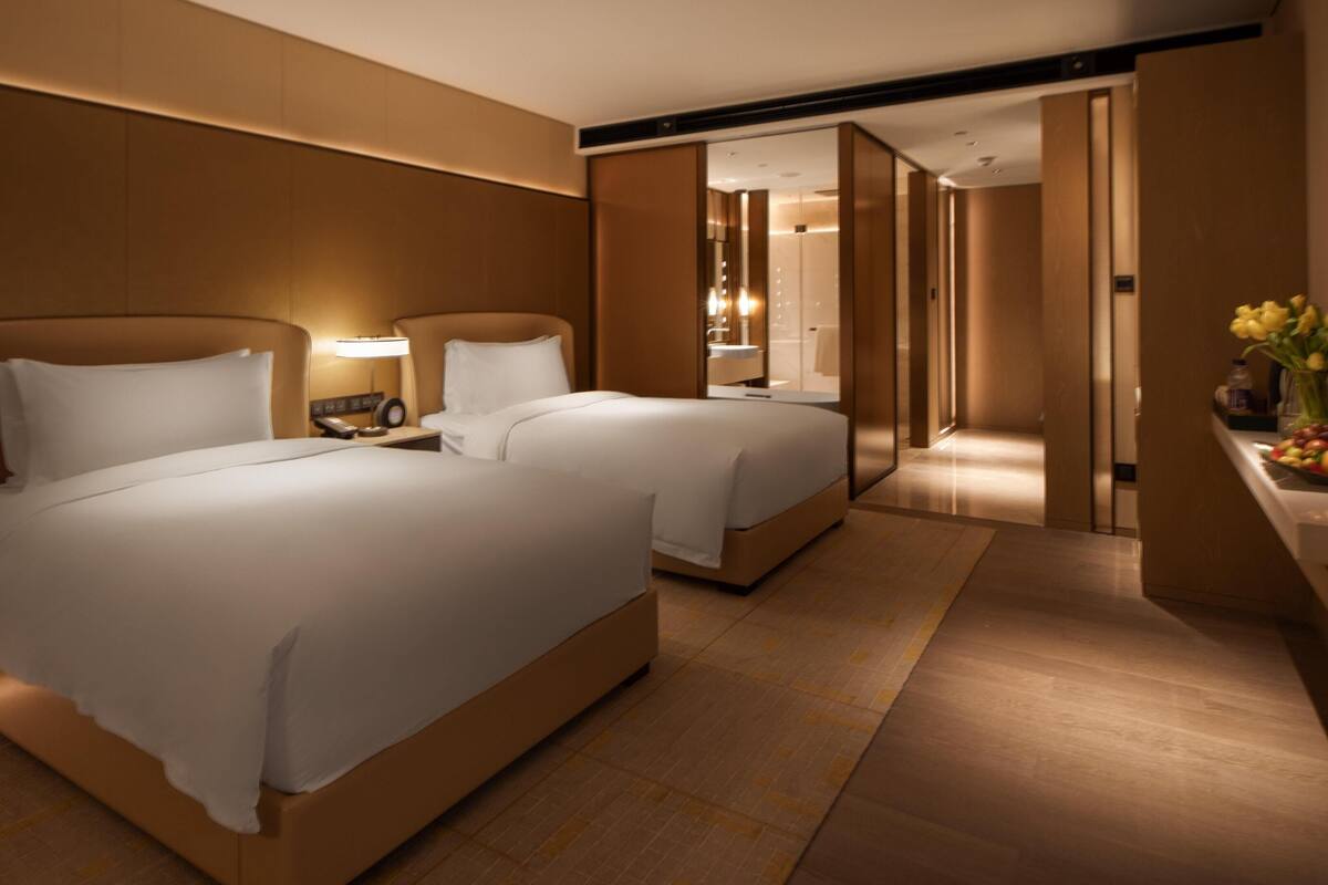 Habitación de hotel con dos camas, una mesita de noche con lámpara, una pequeña mesa con fruta y un espejo.