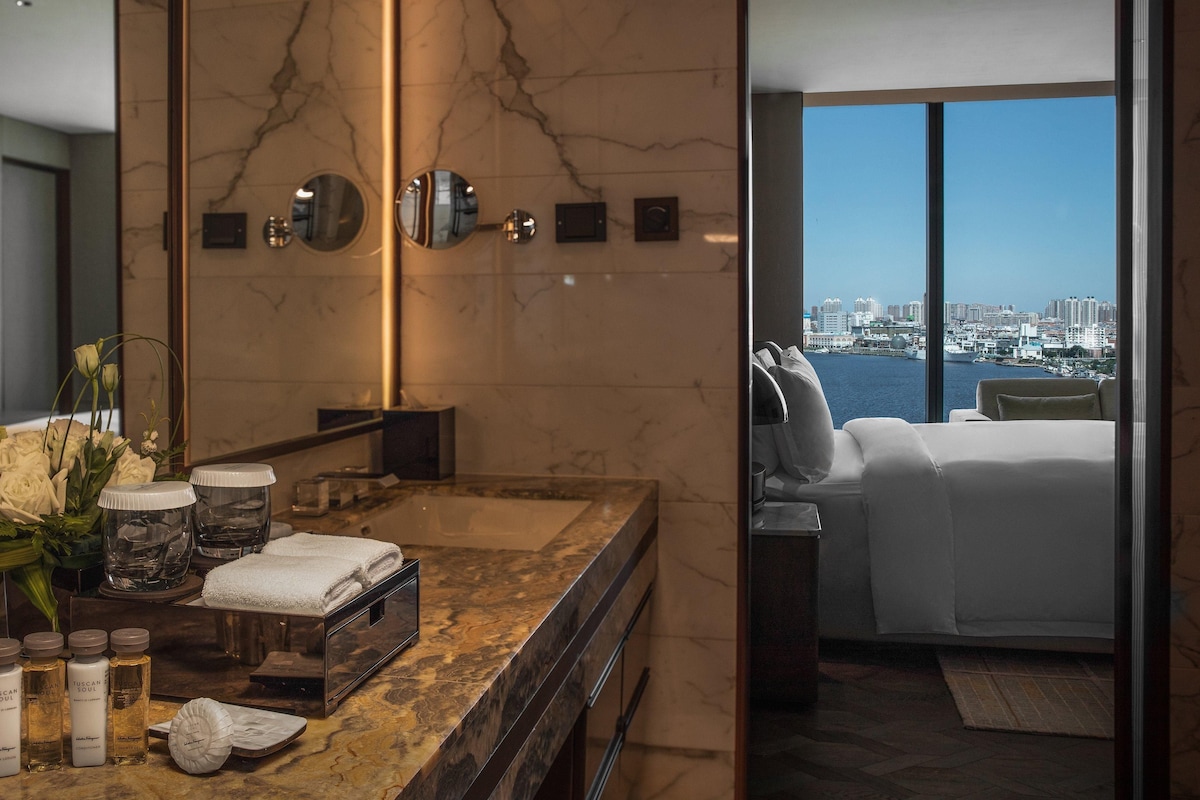 Un baño moderno con encimera de mármol, un espejo grande y vistas de la ciudad a través de un ventanal.