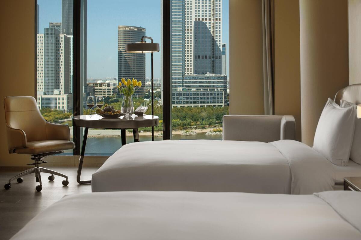 Habitación de hotel con una cama, dos sillas, una mesita con un florero y fruta, una lámpara y vistas a edificios de la ciudad.