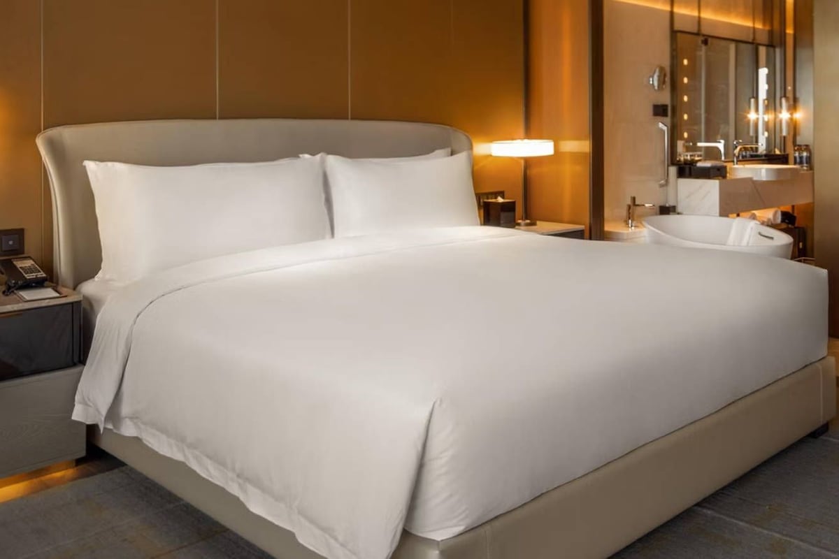 Una cama grande con ropa de cama blanca, una mesita de noche con lámpara y un baño con bañera y espejo.