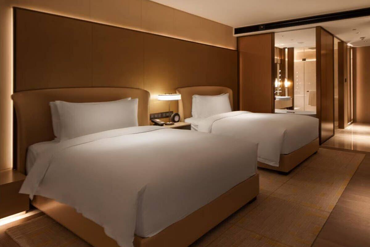 Habitación de hotel con dos camas, una mesita de noche con lámpara y una puerta corredera que da a un baño.