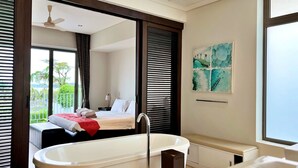 A modern bathroom with a freestanding bathtub, a toilet, and a sink. There is a bed with a red blanket and a view of a balcony with greenery outside. - Baan Yamu By RESAVA (Pa Klok)