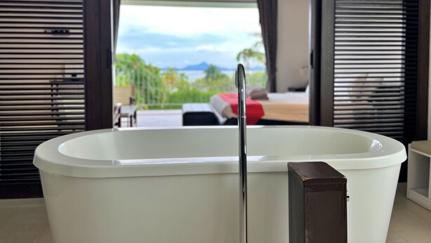 A modern bathroom with a freestanding bathtub, a ceiling fan, and a view of a balcony with outdoor furniture. - Baan Yamu By RESAVA (Pa Klok)