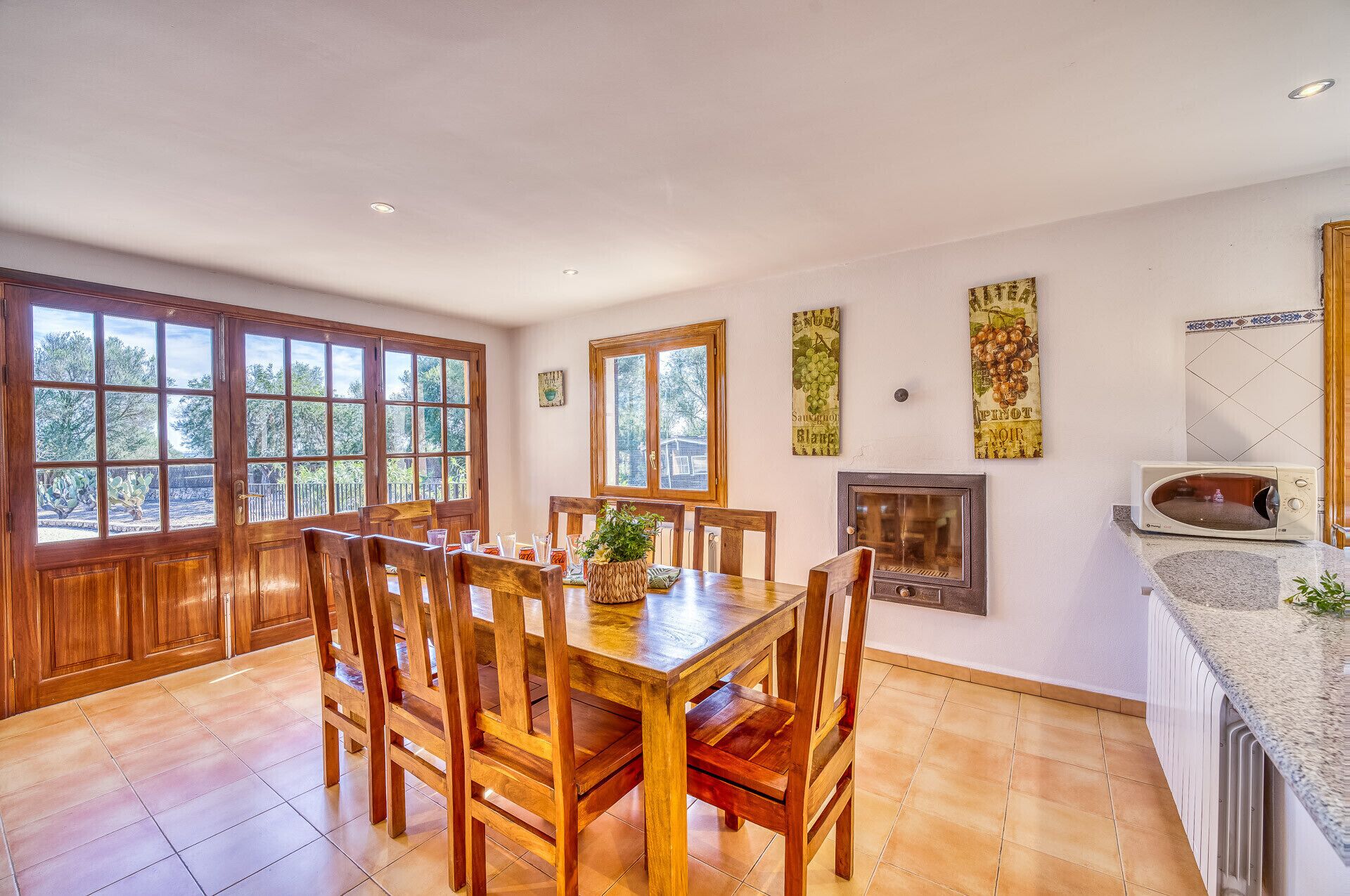 A dining area with a wooden table and chairs, large windows, a fireplace, and kitchen appliances.
