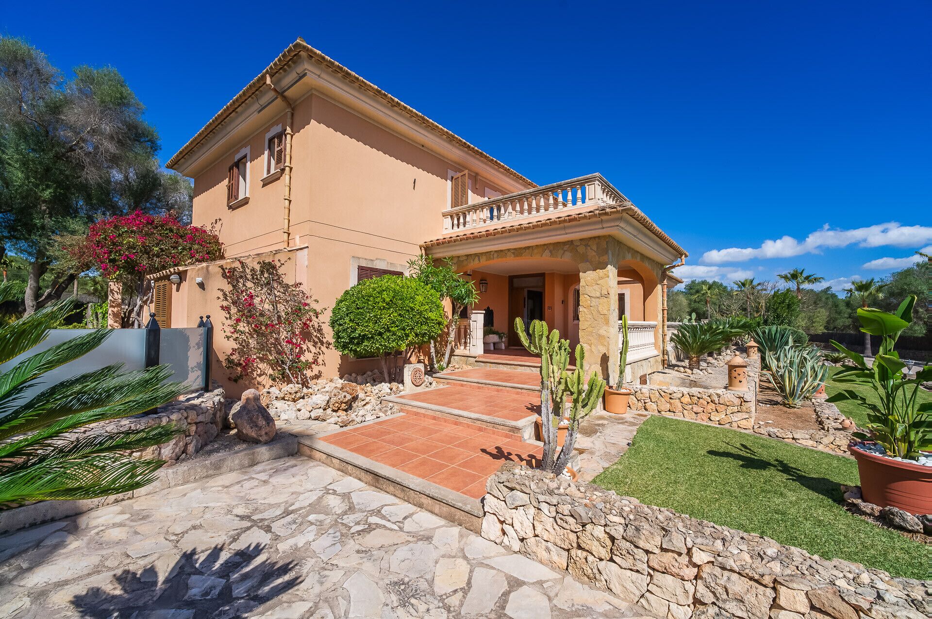 A two-story house with a stone facade, a balcony, and a well-maintained garden with cacti and shrubs.