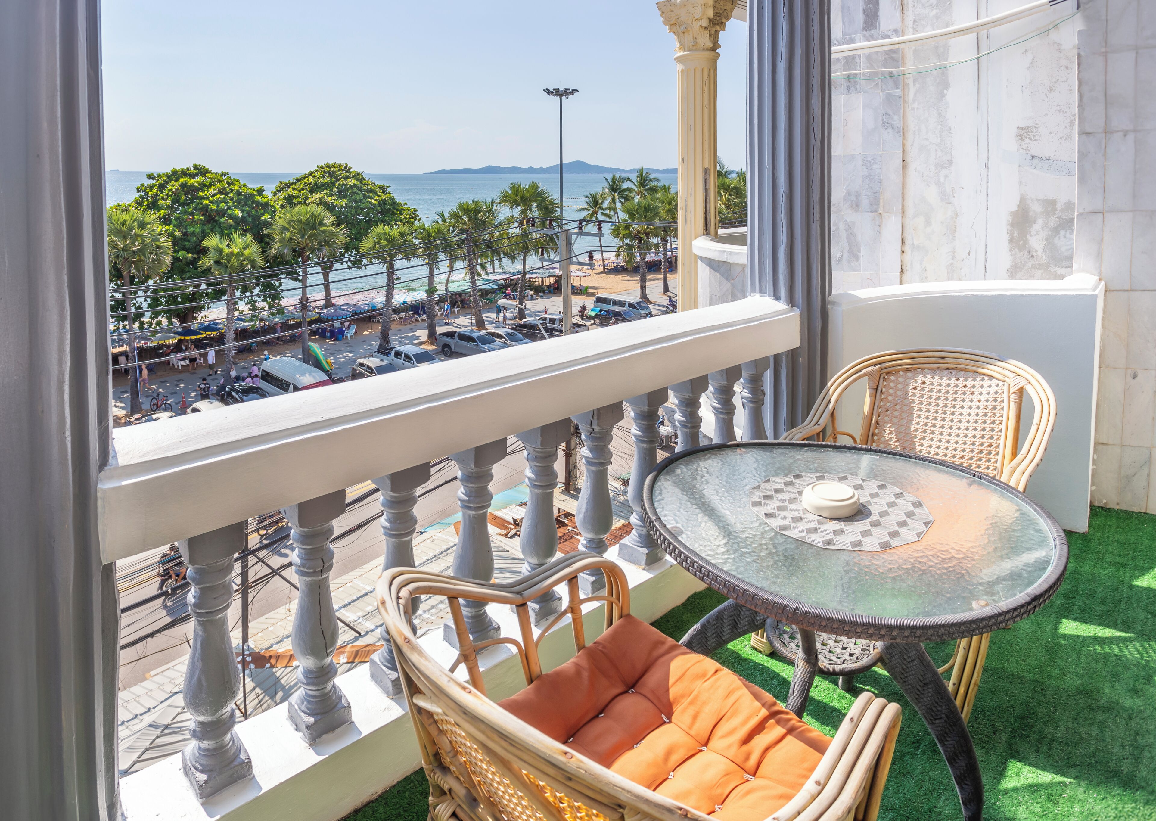 A balcony with wicker chairs and a glass table, overlooking a beach with palm trees and a clear sky.