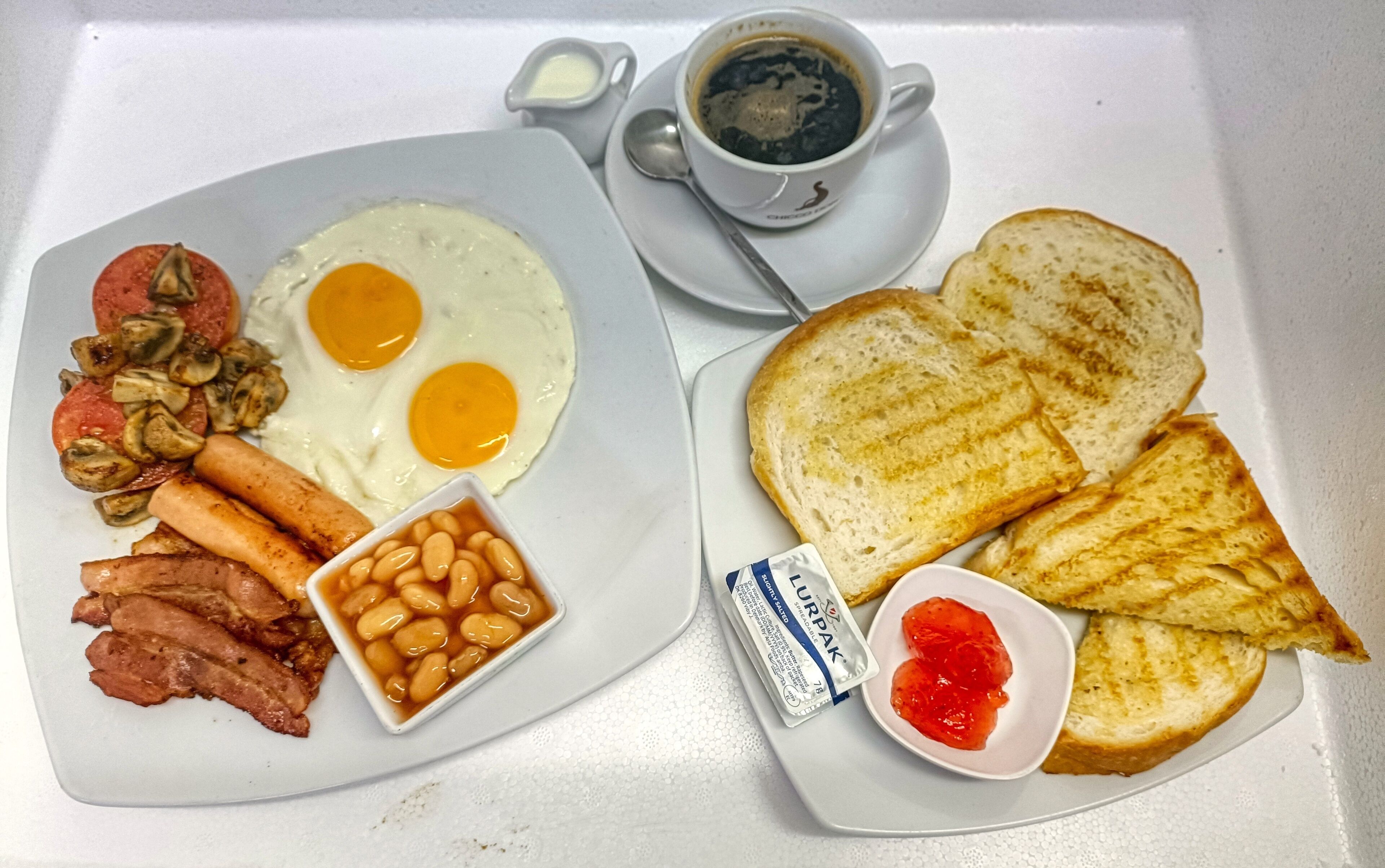 Een ontbijtje met eieren, worstjes, champignons, bonen, toast, jam en een kop koffie.