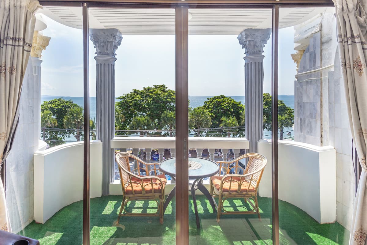 A balcony with a glass door, two wicker chairs, a round table, and a view of the sea and greenery.