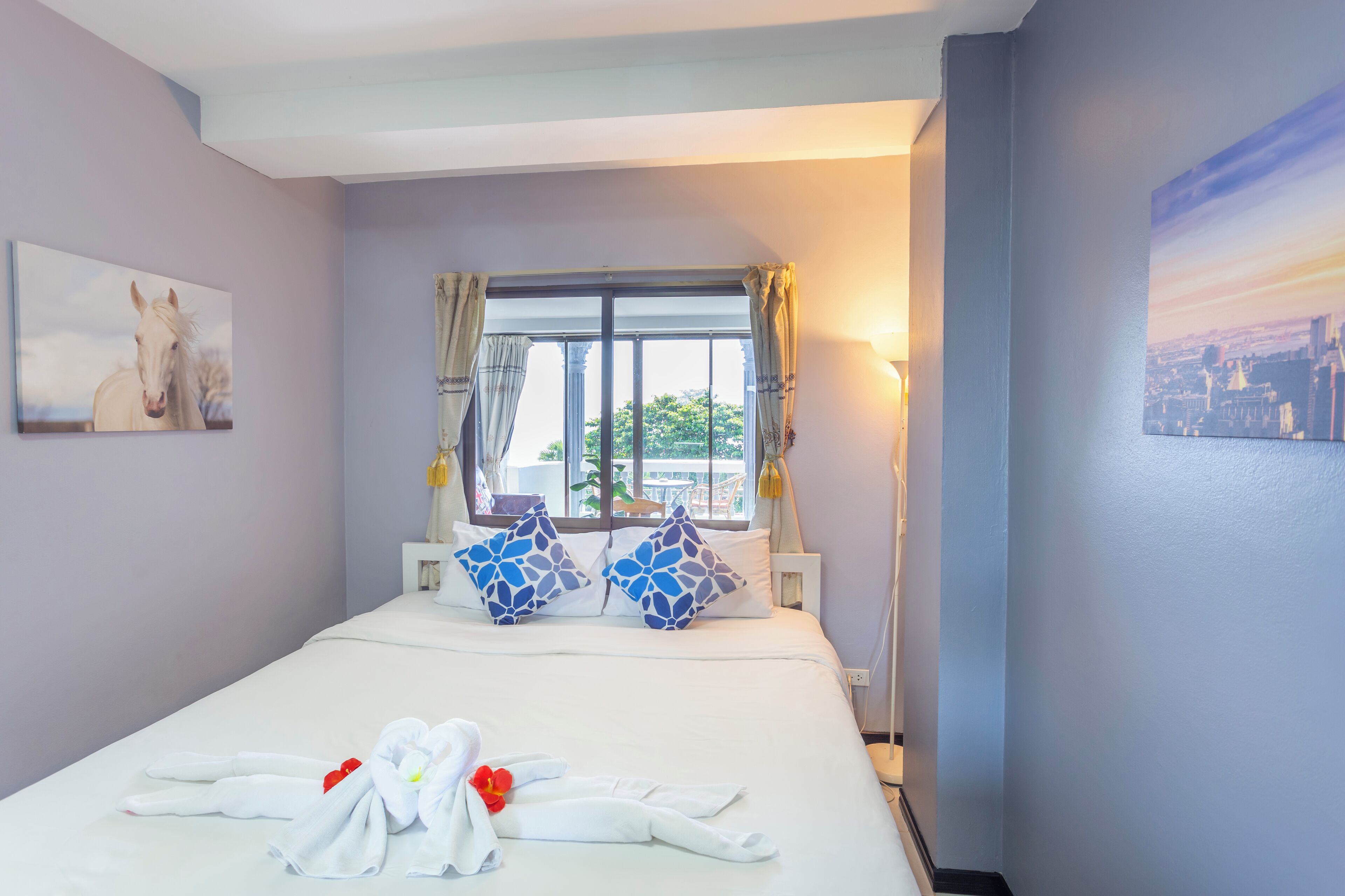 Une chambre d’hôtel avec un lit, une fenêtre avec des rideaux, un tableau représentant un cheval et un tableau représentant un paysage urbain accroché au mur.