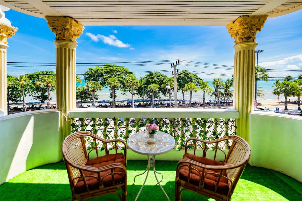 A balcony with two wicker chairs and a small table, overlooking a beach with palm trees and umbrellas.