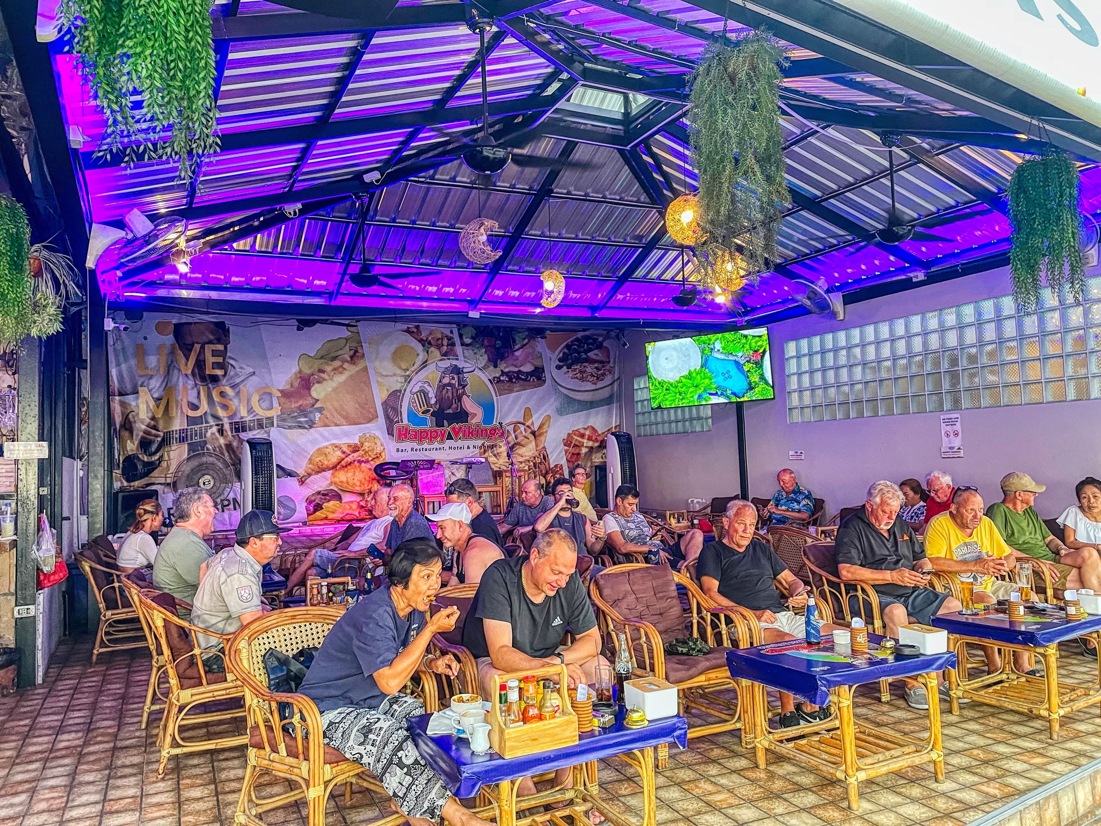 A restaurant with outdoor seating, a large TV screen, and a sign advertising live music.