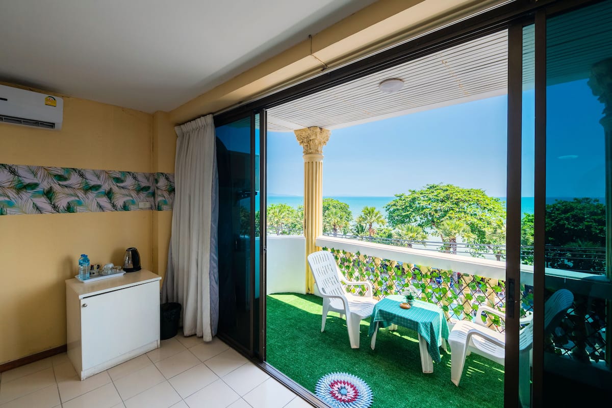 A balcony with a view of the ocean, a small table with a tablecloth, a white chair, and a white cabinet with a water bottle and a kettle.