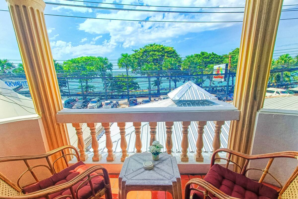 A balcony with wicker chairs and a small table, overlooking a busy street with cars and buildings.