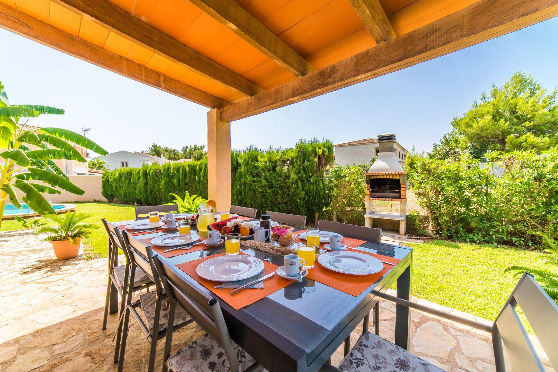 An outdoor dining area with a table set for breakfast, surrounded by greenery and a barbecue grill.