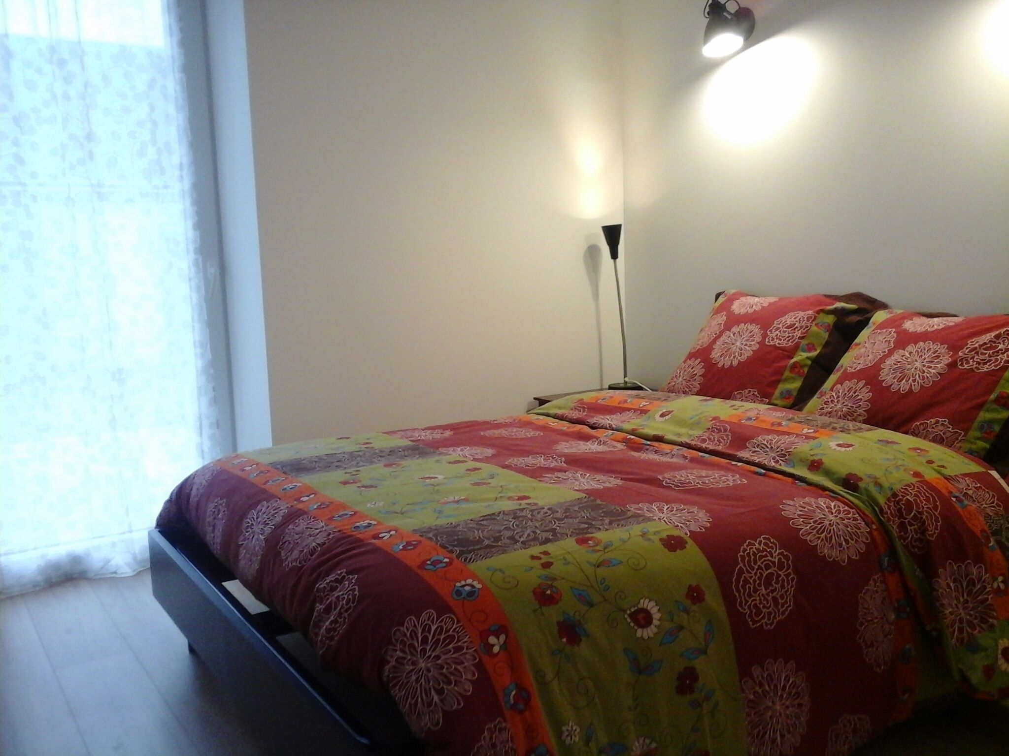 Une chambre à coucher avec un lit recouvert d’une literie aux motifs floraux colorés, une lampe de chevet et une fenêtre ornée de rideaux légers.